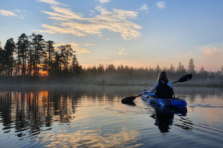 Person kayaking on a calm lake at sunset with trees and cloudy sky reflected on the water.