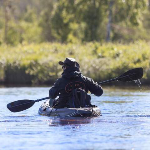 Person kayaking on a calm river near a grassy shore with trees in the background.