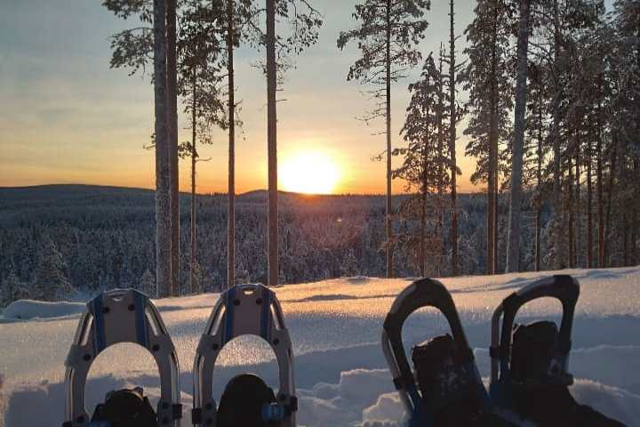 Person wearing snowshoes sitting, watching a sunset over a snowy forest.