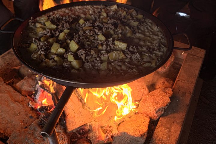 Large pan of food cooking over open fire with rocks and people around.