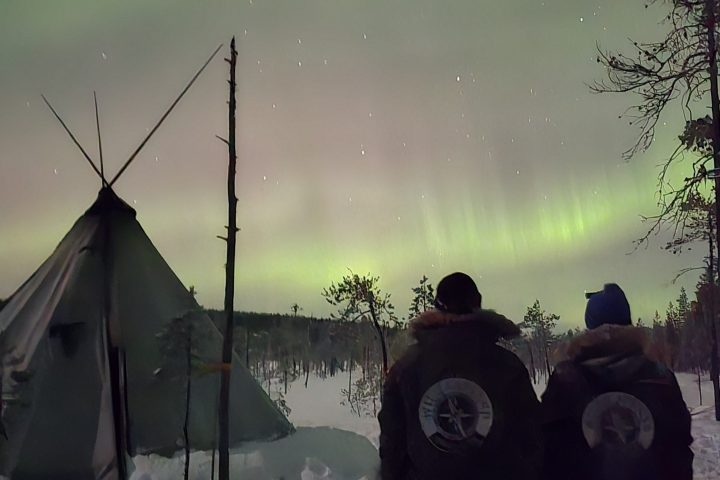 People in winter gear watching northern lights near a tent and snowy landscape.