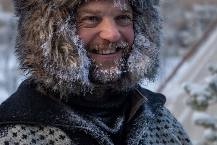 Person in fur hat and frosty beard, wearing patterned sweater outdoors.